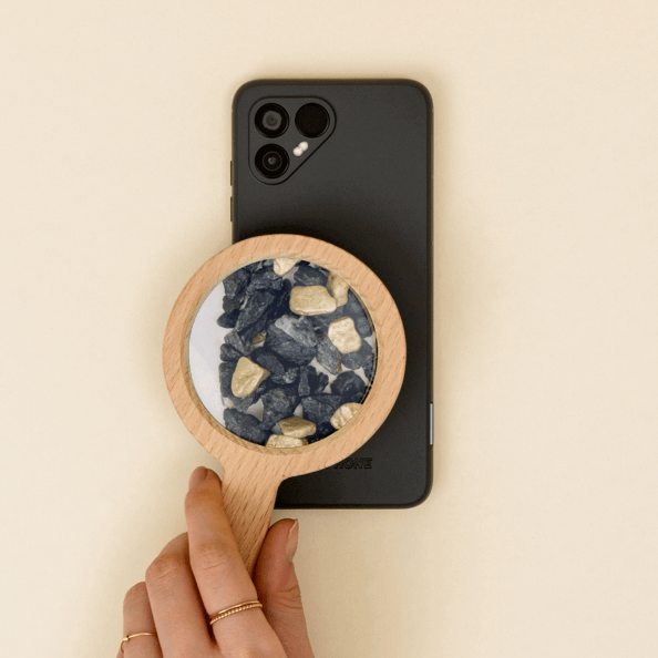 Creative presentation of a black Fairphone with a wooden-handled magnifying glass revealing a rocky texture beneath